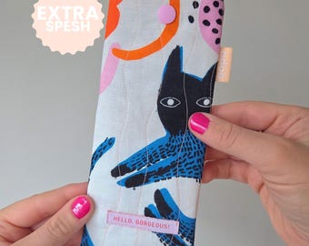 May include: A white quilted glasses case with a colorful cat design. The case features a black cat with blue accents, orange and pink abstract shapes, and the text "HELLO, GORGEOUS!" on a pink label.