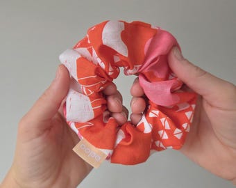 May include: A colorful scrunchie with a vibrant pattern of orange, white, and pink shapes. The scrunchie is held between two hands, showcasing its design and texture. A small tag with the text "aopa" is visible.