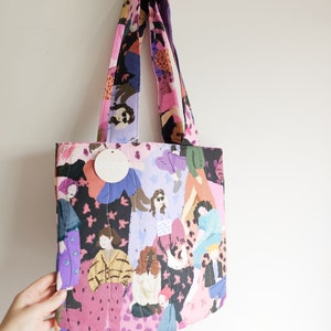 Pu&ograve; includere: Una borsa tote trapuntata con una stampa colorata raffigurante figure in vari outfit. La borsa ha cinghie rosa e viola e un'etichetta con la scritta "pops". La borsa &egrave; appesa a una parete bianca.