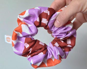 May include: A colorful scrunchie with a vibrant pattern of orange, purple, white, and maroon geometric shapes. The hair accessory is held by a hand and features a small white tag with the word "copa".