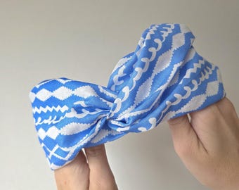 May include: A blue and white patterned headband. The fabric is a vibrant blue with a repeating white design of wavy lines and diamond shapes. The headband is twisted at the top, creating a stylish accessory.