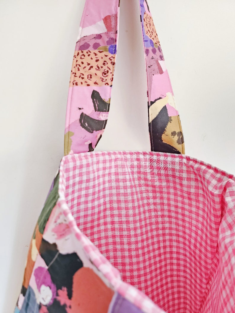 Pu&ograve; includere: Una borsa tote con esterno rosa fantasia e interno a quadretti rosa. L'esterno presenta un collage di forme astratte in rosa, viola, nero e beige. I manici sono rosa e fantasia, in linea con il design esterno.
