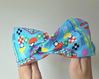 May include: A blue headband with a colorful geometric pattern. The pattern includes shapes in red, orange, yellow, and green. The headband is held up by two hands.