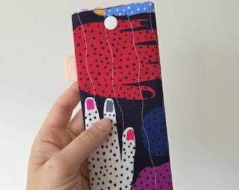 May include: A rectangular fabric case with a colorful abstract design. The case features a hand-shaped pattern with dotted details and painted fingernails. A white button secures the case, and a small tag with the word "bopa" is visible.