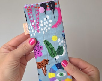 May include: A rectangular, fabric glasses case with a light blue background and a colorful jungle-themed pattern. The pattern includes a leopard, flowers, and an eye design. The case has a pink button closure and a small tag that says "odoo".