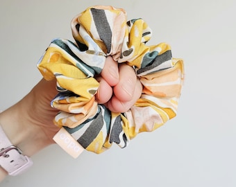 Può includere: Un colorato elastico per capelli con un vivace motivo astratto in giallo, grigio, pesca e verde acqua. L'elastico è tenuto in mano e una piccola etichetta con la parola "oopa" è visibile. Questo accessorio per capelli è perfetto.