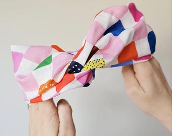 May include: A colorful fabric headband with a knotted design. The headband features a pattern of pink, blue, orange, green, and yellow squares on a white background. The accessory is held by two hands against a neutral background.