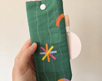 May include: A green fabric glasses case with a white button closure. The case features a stitched design with vertical lines and colorful geometric shapes in orange, yellow, and pink. A tag with the word "indoo" is attached.