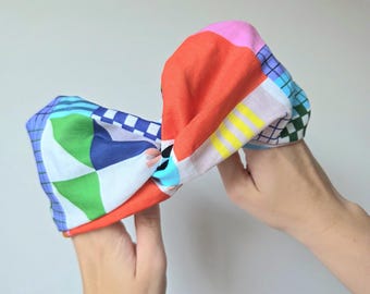 Può includere: Una fascia colorata con un design annodato. La fascia presenta un vivace mix di motivi geometrici e colori a tinta unita, tra cui rosso, rosa, blu, verde e giallo. L'accessorio è tenuto in alto da due mani.