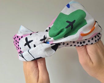May include: A white headband with a colorful abstract pattern. The design includes green, pink, black, and orange shapes, along with black cross marks and small black dots. The headband is held up by two hands.