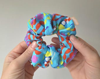 May include: A colorful scrunchie with a vibrant pattern of red, pink, yellow, and white shapes on a light blue background. The scrunchie is held between two hands. A small tag with the word "odoo" is visible.