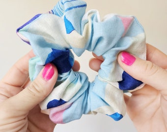 May include: A light blue scrunchie with a colorful abstract pattern of blue, pink, and white shapes. The scrunchie is held between two hands with pink nail polish.