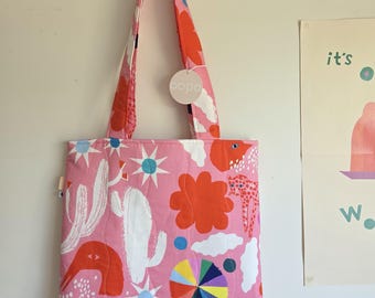 May include: Pink tote bag with a colorful pattern featuring cacti, flowers, and abstract shapes. The bag has matching straps and a tag that says "bopa". The bag is hanging on a white wall.