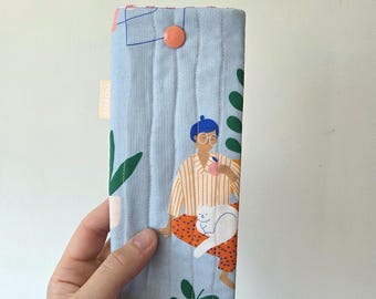 May include: A light blue quilted fabric case with a button closure. The case features a printed illustration of a person with a cat, drinking from a mug. Green leaves and geometric shapes are also printed on the case.