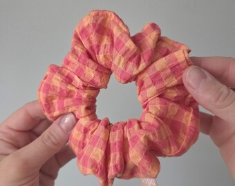 May include: A fabric hair scrunchie featuring a vibrant gingham check pattern in shades of bright pink and soft orange. The accessory has a gathered, ruffled texture and includes a small, rectangular fabric tag attached to the outer edge.