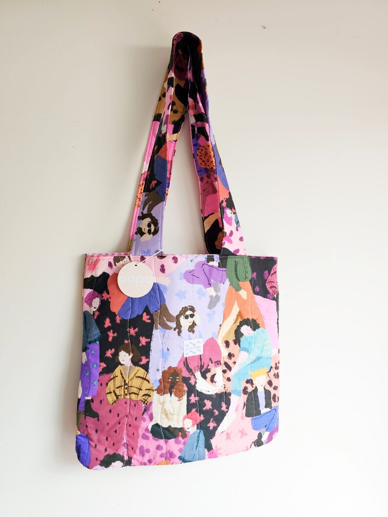 Pu&ograve; includere: Una borsa tote trapuntata con un motivo colorato di figure in varie pose. La borsa presenta un mix di tonalit&agrave; rosa, viola, nere e gialle. I manici e il corpo della borsa sono realizzati con lo stesso tessuto fantasia. &Egrave; attaccato un piccolo tag rotondo con la scritta "oops".