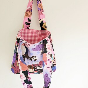 Pu&ograve; includere: Una borsa tote con un motivo astratto rosa, viola e nero. La borsa ha lunghe cinghie e un interno a quadretti vichy. L'esterno presenta un design ripetuto di figure e forme. La borsa &egrave; appesa su uno sfondo bianco.