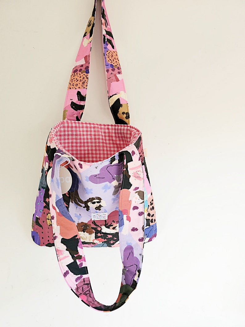 Pu&ograve; includere: Una borsa tote con un motivo astratto rosa, viola e nero. La borsa ha lunghe cinghie e un interno a quadretti vichy. L'esterno presenta un design ripetuto di figure e forme. La borsa &egrave; appesa su uno sfondo bianco.