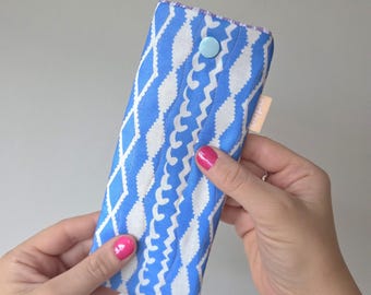 May include: A blue and white patterned glasses case with a button closure. The case features a repeating diamond and wavy design. The case is held by a person with pink nail polish.