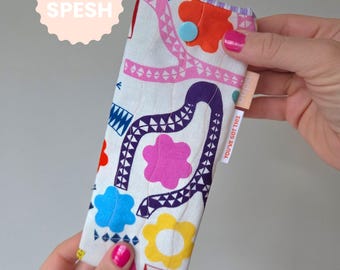 May include: A white fabric glasses case with a colorful floral pattern, featuring pink, blue, yellow, and purple shapes. The case has a blue button closure and a small tag that says "YOU'VE GOT THIS". The text "EXTRA SPESH" is on a pink circle.