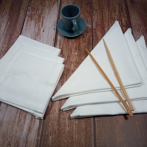 Può includere: Un set di quattro tovaglioli di lino bianco piegati a forma di triangolo, con due bacchette di legno sopra. I tovaglioli sono disposti su una superficie di legno, con una tazza e un piattino in ceramica blu sullo sfondo.