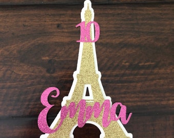 May include: Gold glitter Eiffel Tower cake topper with a pink glitter "10" and "Emma" written on the front.