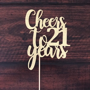 May include: Gold glitter cake topper with the text "Cheers to 21 years".