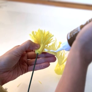 May include: A hand holds a yellow paper flower with a green wire stem. White glue is being applied to the base of the flower.