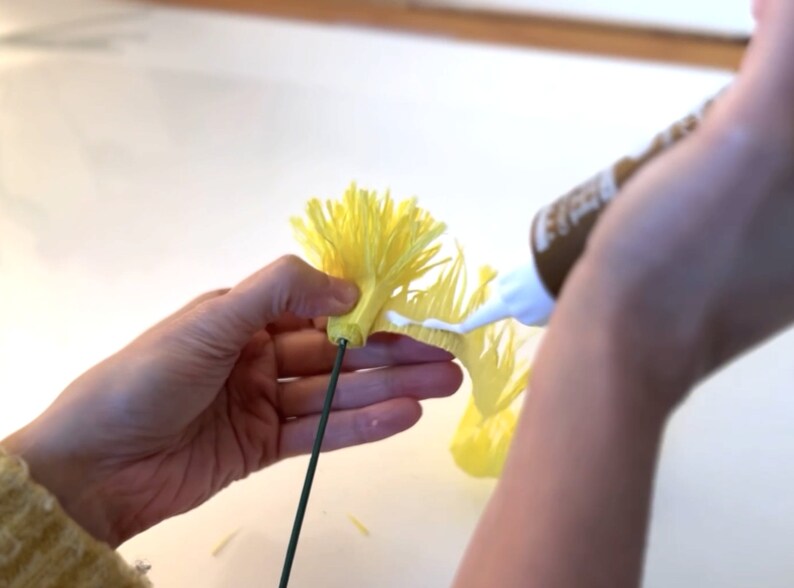May include: A hand holds a yellow paper flower with a green wire stem. White glue is being applied to the base of the flower.