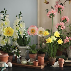 May include: A collection of paper flowers in various colors, including yellow, pink, and white, arranged in terracotta pots and a white ceramic planter. The flowers are displayed on a wooden table with a brown surface.