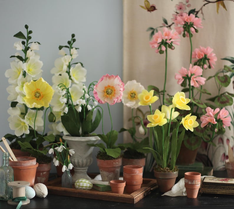 May include: A collection of paper flowers in various colors, including yellow, pink, and white, arranged in terracotta pots and a white ceramic planter. The flowers are displayed on a wooden table with a brown surface.