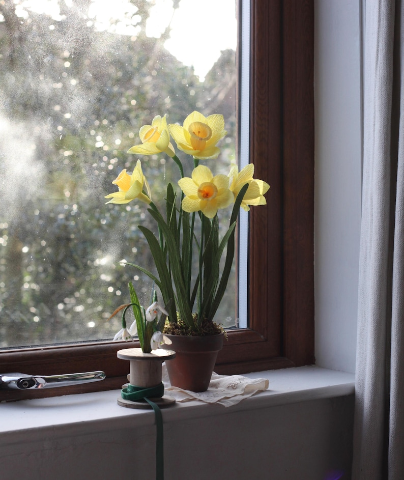 May include: A bouquet of yellow daffodils in a terracotta pot sits on a windowsill. The flowers are in full bloom and the pot is surrounded by green leaves. The window is open and there is a view of a blurry green background.