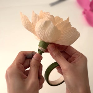 May include: A hand is wrapping green floral tape around the base of a pale peach paper flower. The tape is being held in place with a green floral tape dispenser.