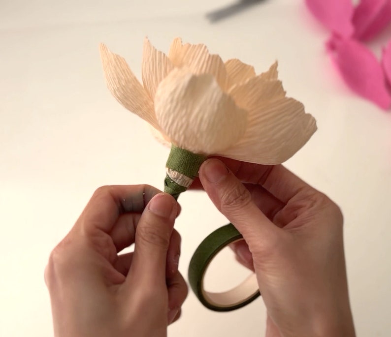 May include: A hand is wrapping green floral tape around the base of a pale peach paper flower. The tape is being held in place with a green floral tape dispenser.