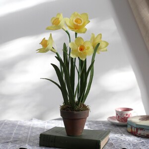 May include: A potted arrangement of yellow paper daffodils in a terracotta pot. The pot is sitting on top of a vintage book.