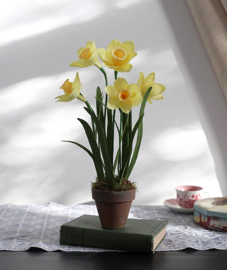 May include: A potted arrangement of yellow paper daffodils in a terracotta pot. The pot is sitting on top of a vintage book.
