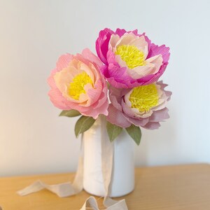 May include: A bouquet of three pink and yellow paper peonies in a white vase with a beige ribbon.