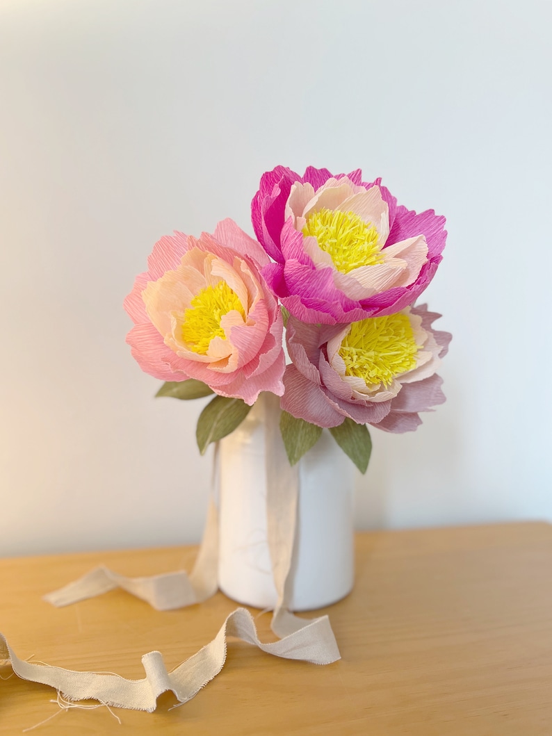 May include: A bouquet of three pink and yellow paper peonies in a white vase with a beige ribbon.