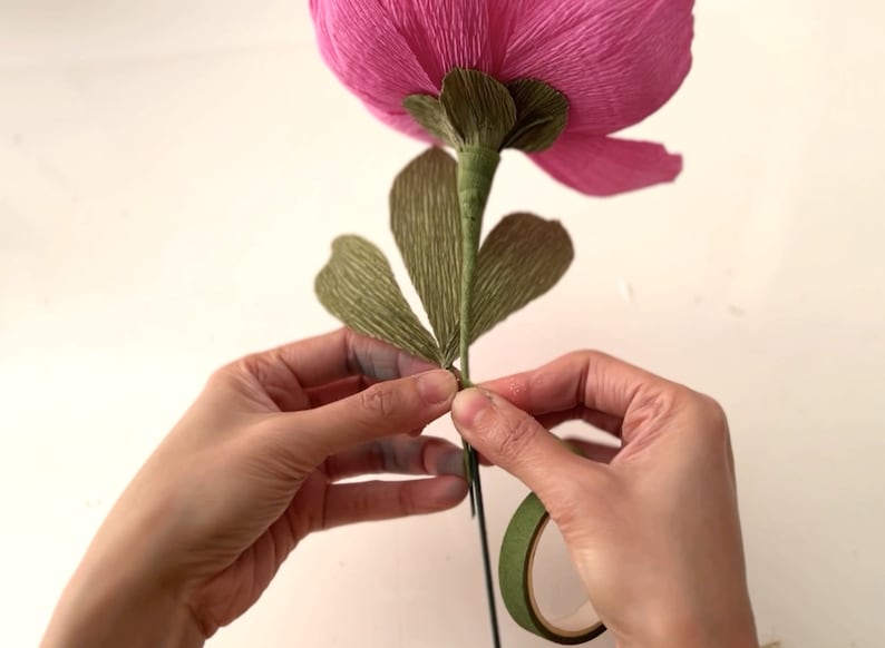 May include: A hand is holding a pink paper flower with green paper leaves and securing the leaves to the stem with green floral tape.
