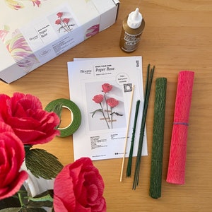 May include: A paper rose craft kit with instructions, tools, and materials. The kit includes green floral tape, green and brown floral wire, a wooden skewer, and red crepe paper. The kit is packaged in a white box with the text "Make Your Own Paper Rose" and "Blooms by Dream".