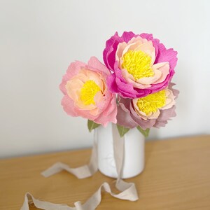May include: A bouquet of three paper peonies in shades of pink and yellow, arranged in a white vase. The vase is sitting on a wooden table with a beige ribbon.
