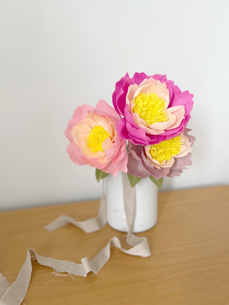 May include: A bouquet of three paper peonies in shades of pink and yellow, arranged in a white vase. The vase is sitting on a wooden table with a beige ribbon.