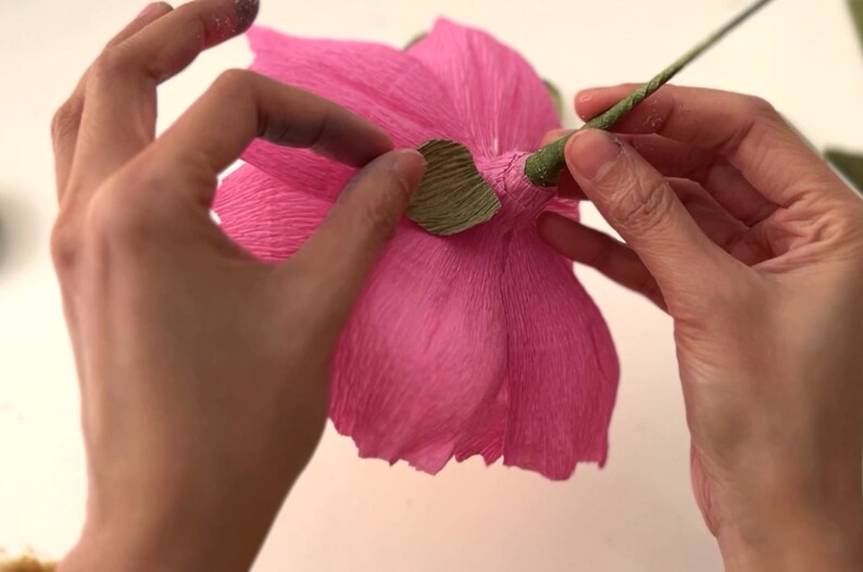 May include: A hand is holding a pink paper flower and attaching a green paper leaf to the base of the flower.