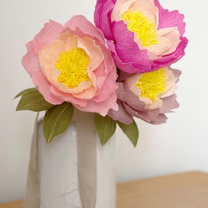 May include: A bouquet of three paper peonies in shades of pink and yellow, arranged in a white vase with a beige ribbon.