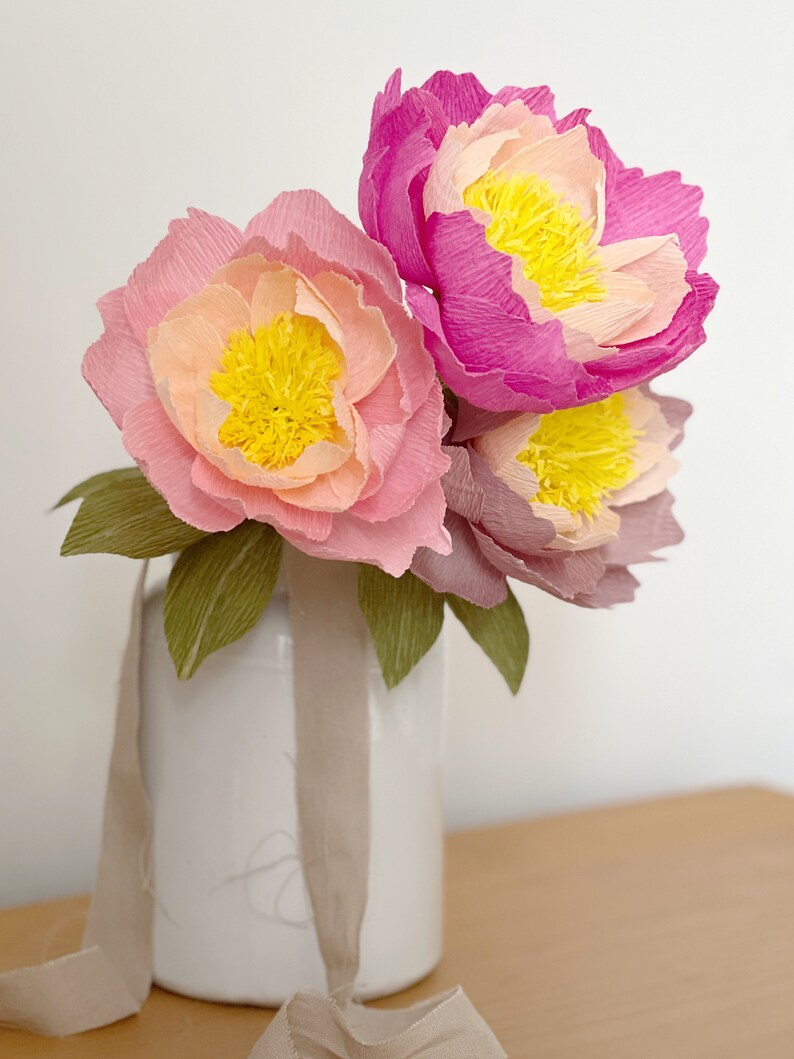 May include: A bouquet of three paper peonies in shades of pink and yellow, arranged in a white vase with a beige ribbon.