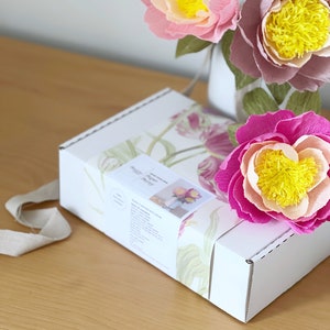 May include: A white box with a floral design on the front. The box is labeled "Paper Peony". Three paper peonies in pink, peach, and purple are arranged in a vase behind the box.
