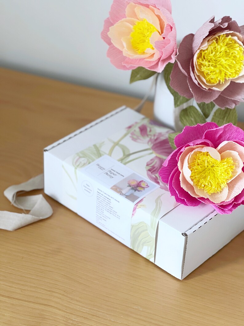 May include: A white box with a floral design on the front. The box is labeled "Paper Peony". Three paper peonies in pink, peach, and purple are arranged in a vase behind the box.