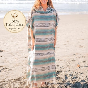 Puede incluir: Una mujer con un poncho de playa largo y a rayas con capucha, de pie en una playa de arena. El poncho presenta rayas horizontales en tonos azules, beige y blanco. El texto "100% Turkish Cotton" es visible.