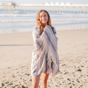 May include: A young woman standing on a sandy beach, wrapped in a striped pink and white beach towel. She is smiling and looking at the camera.