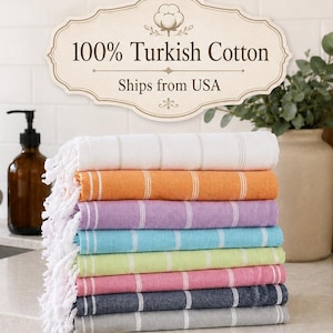 May include: A stack of colorful Turkish cotton towels in various shades, including white, orange, purple, blue, green, pink, navy, and gray. Each towel has a striped design and fringed edges. The towels are displayed on a countertop.